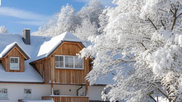 Uma casa de madeira de dois andares com telhado coberto de neve e árvores ao redor, com a inscrição
