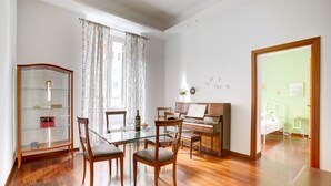 A room with a piano, a glass table with chairs, a bookshelf, and a bed with a nightstand.