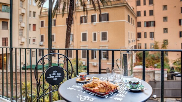 A balcony with a table set for a meal, featuring croissants and a pitcher of water, overlooking a cityscape with palm trees and buildings.