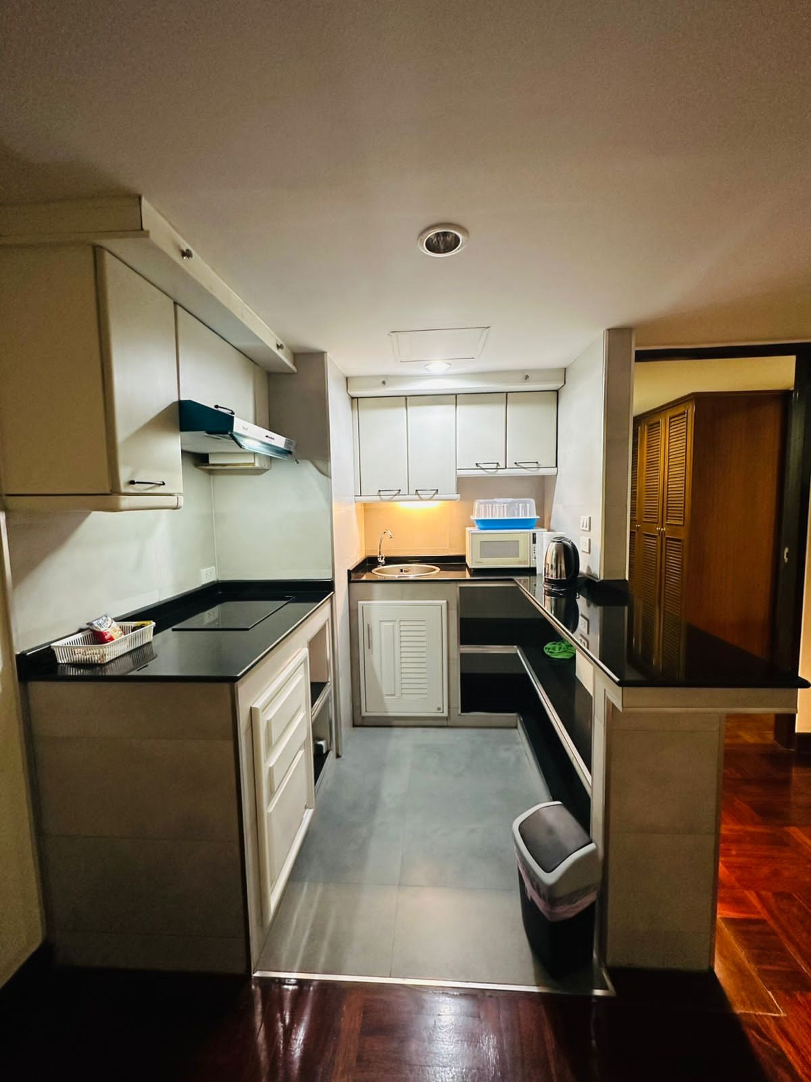 A compact kitchen with white cabinets, a black countertop, a sink, a microwave, and a trash bin.