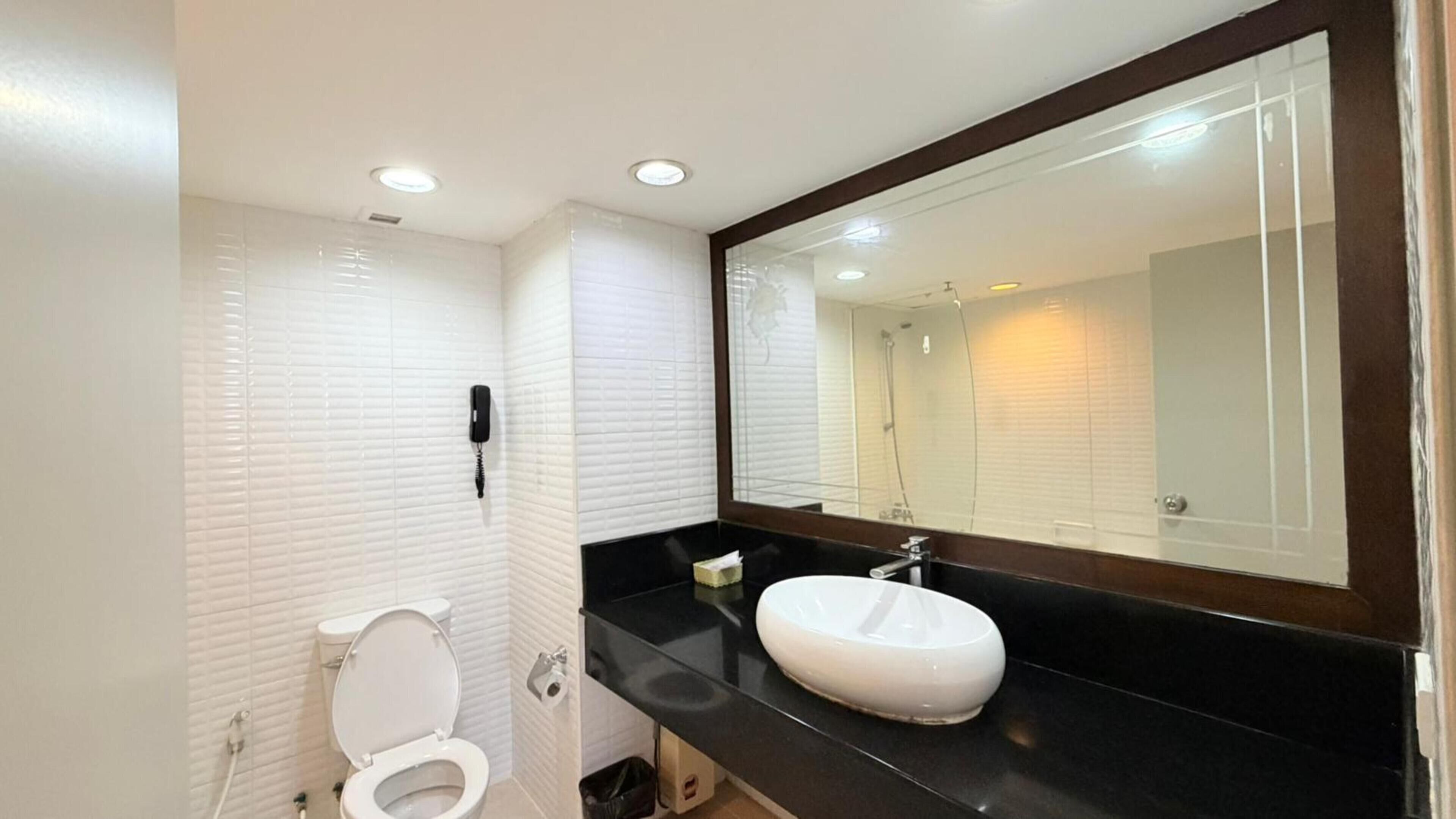 Une salle de bain moderne avec des murs recouverts de carreaux blancs, des toilettes blanches, un comptoir noir, un lavabo blanc et un grand miroir.