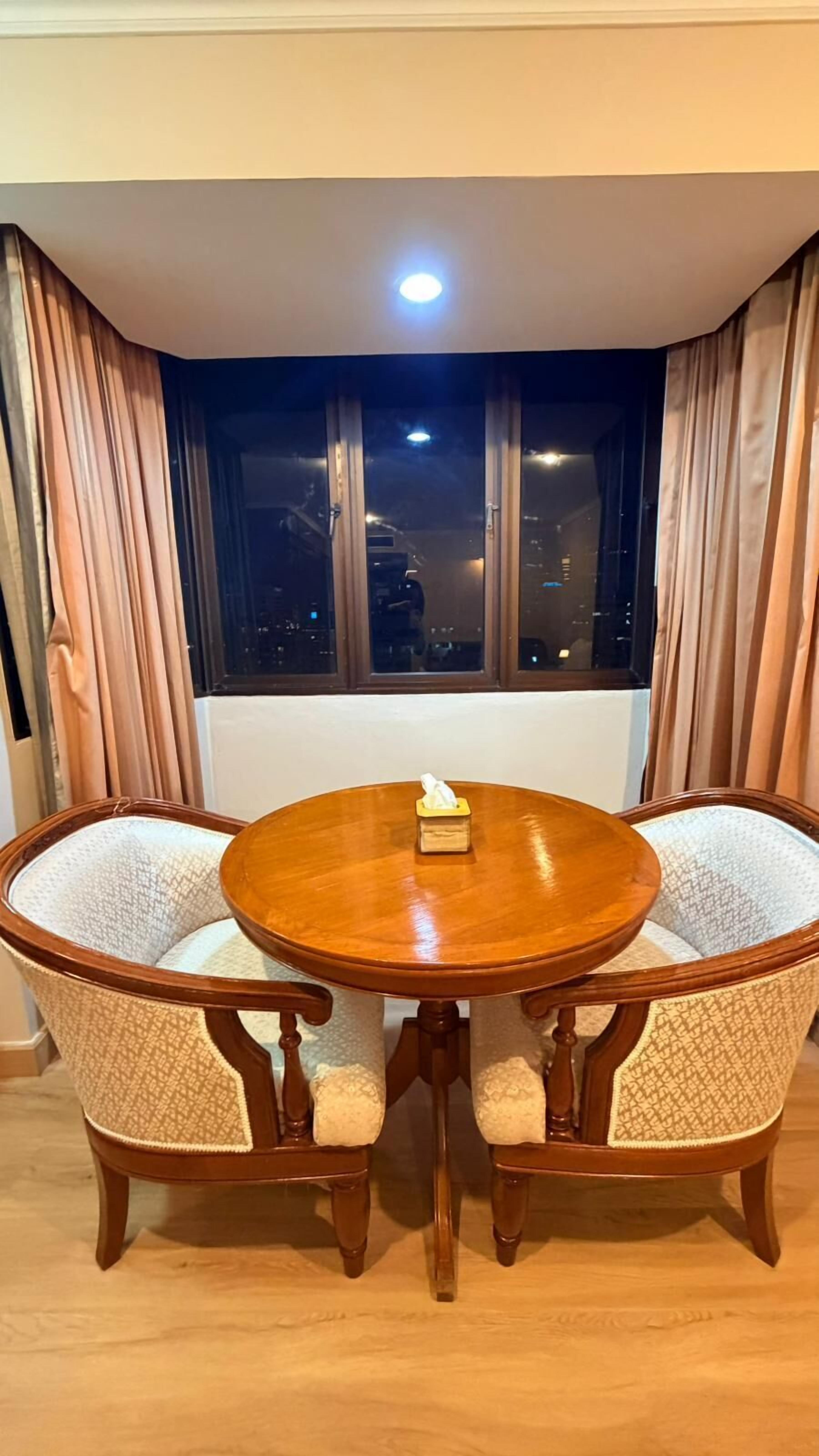Een ronde houten eettafel met twee gestoffeerde stoelen, een zakdoos op de tafel en een uitzicht op de stad door een raam met gordijnen.