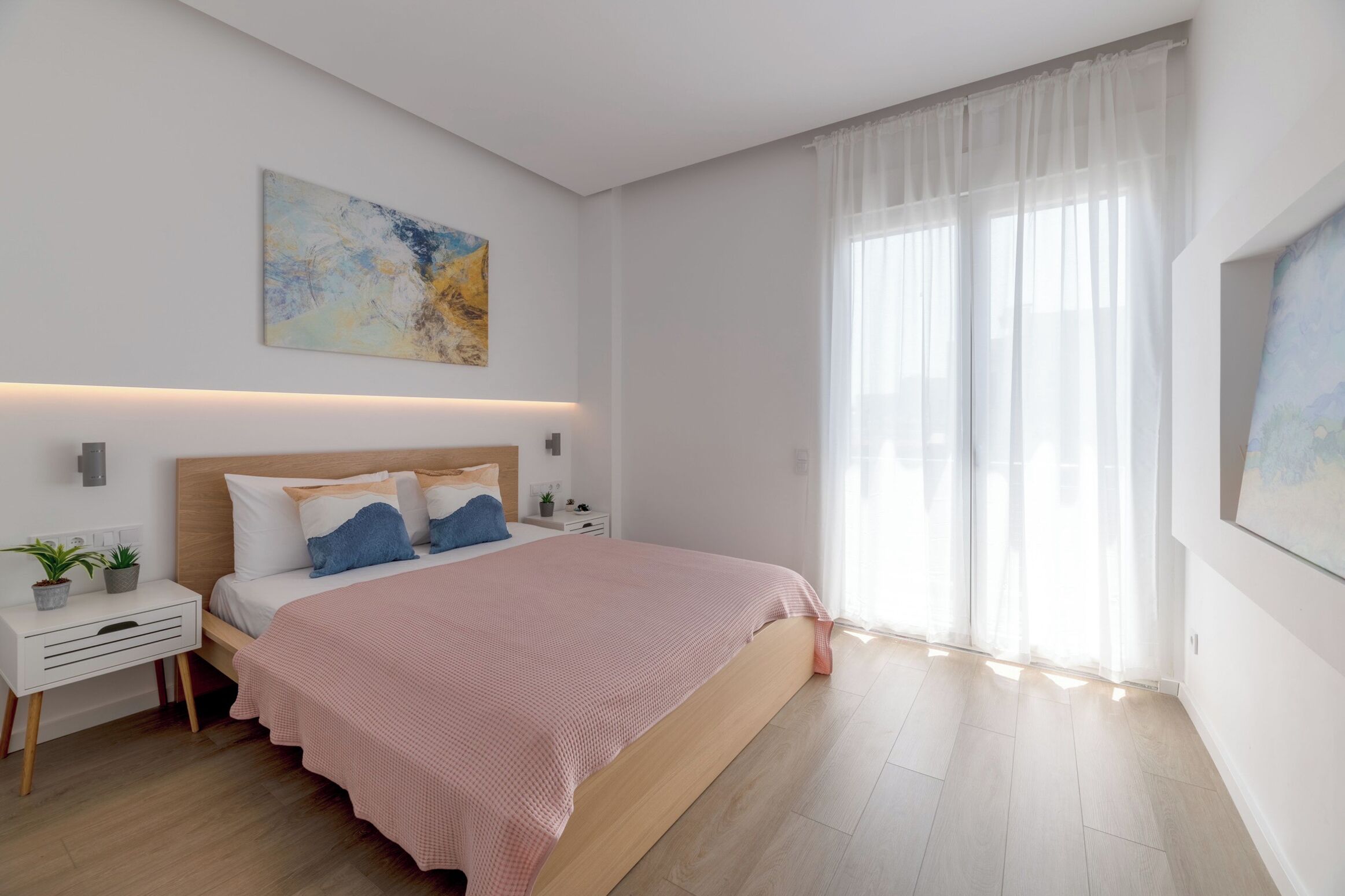 A modern bedroom with a large bed, bedside tables, a nightstand, and a window with sheer curtains.