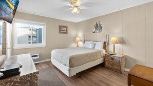 A bedroom with a bed, bedside lamps, a ceiling fan, a window with a view of buildings, and a small desk with a computer. - A Place at the Beach III 9520-2B Summer Escape at Myrtle Beach Ocean View Condo (Myrtle Beach)