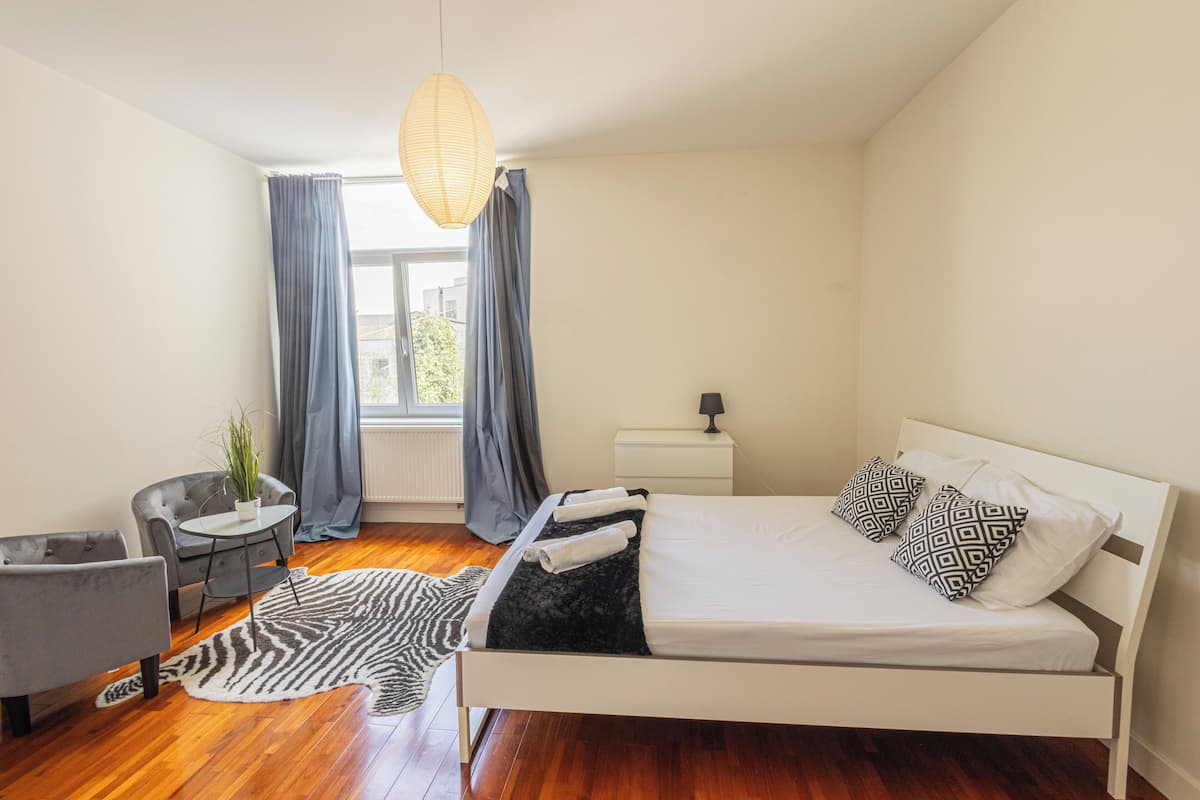 Un dormitorio con cama, una silla, una mesita, una lámpara, una planta y una ventana con cortinas.
