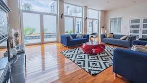 A spacious living room with large windows, two blue sofas, a red bean bag, and a patterned rug. - Chambres Cosy dans Une Grande maison (Brussels)