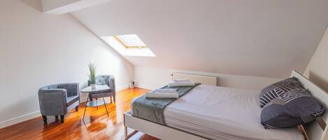 A loft-style bedroom with a sloped ceiling, a skylight, a bed with pillows, a chair, and a small table.
