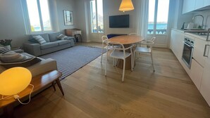 A modern living room with a sofa, dining table, and chairs, a kitchen with white cabinets, and large windows offering a view of the sea. - Beccherie 90 (Intra)