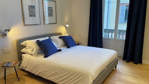 A neatly made bed with white and blue bedding, two framed pictures on the wall, a small table with a magazine, and a window with curtains. - Beccherie 90 (Intra)