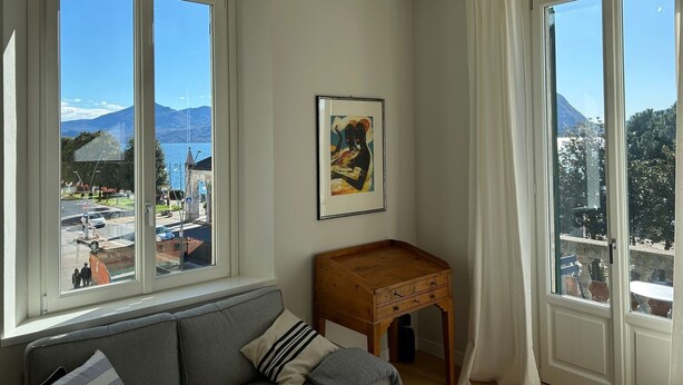 A room with a sofa, a wooden side table, and a framed picture on the wall. There are two windows with a view of a lake and mountains. - Beccherie 90 (Intra)