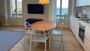 A modern kitchen with a dining area, a TV mounted on the wall, and large windows overlooking a scenic view. - Beccherie 90 (Intra)