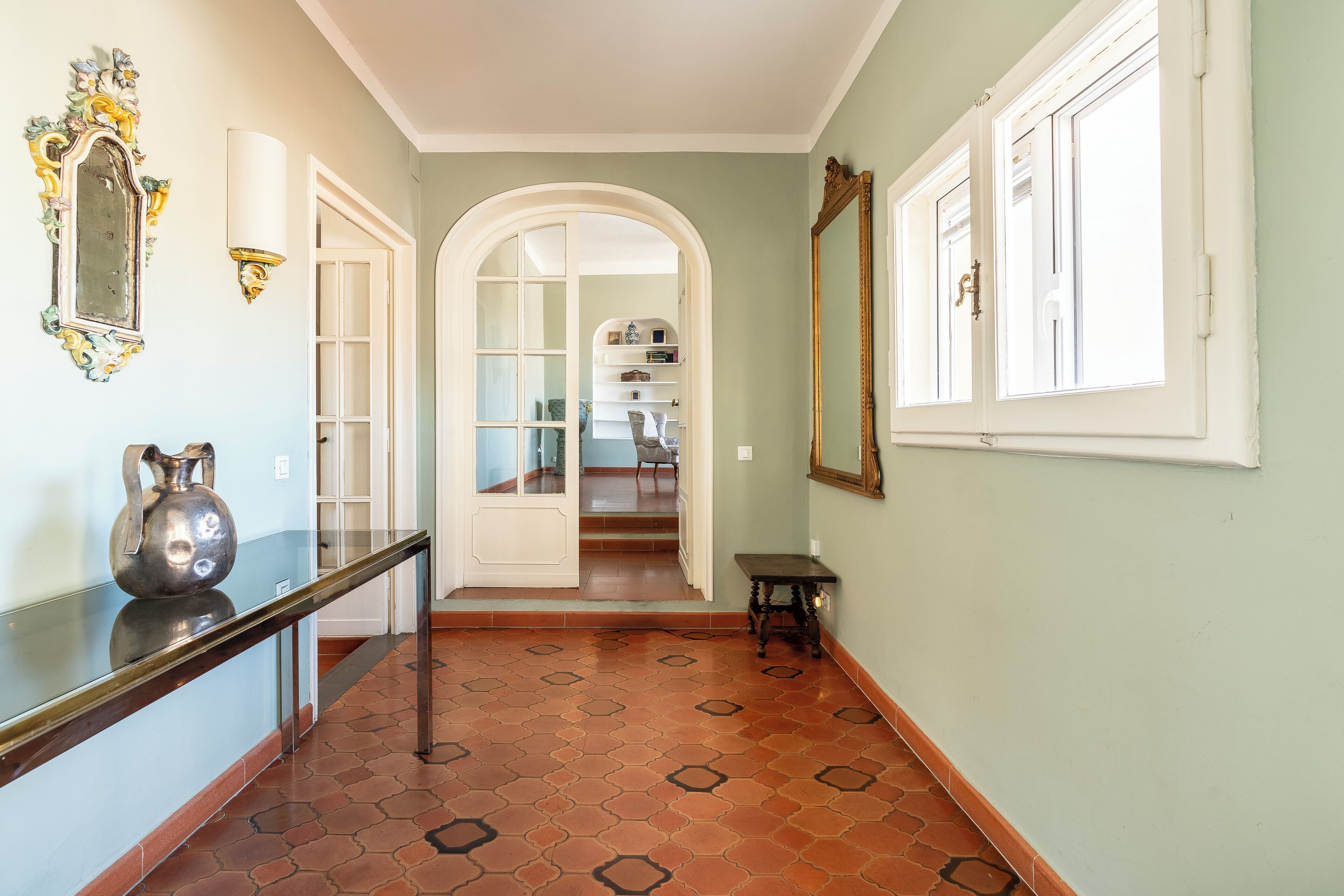 A hallway with a tiled floor, a glass display case, a metal vase, a white door, and a staircase leading to another room.