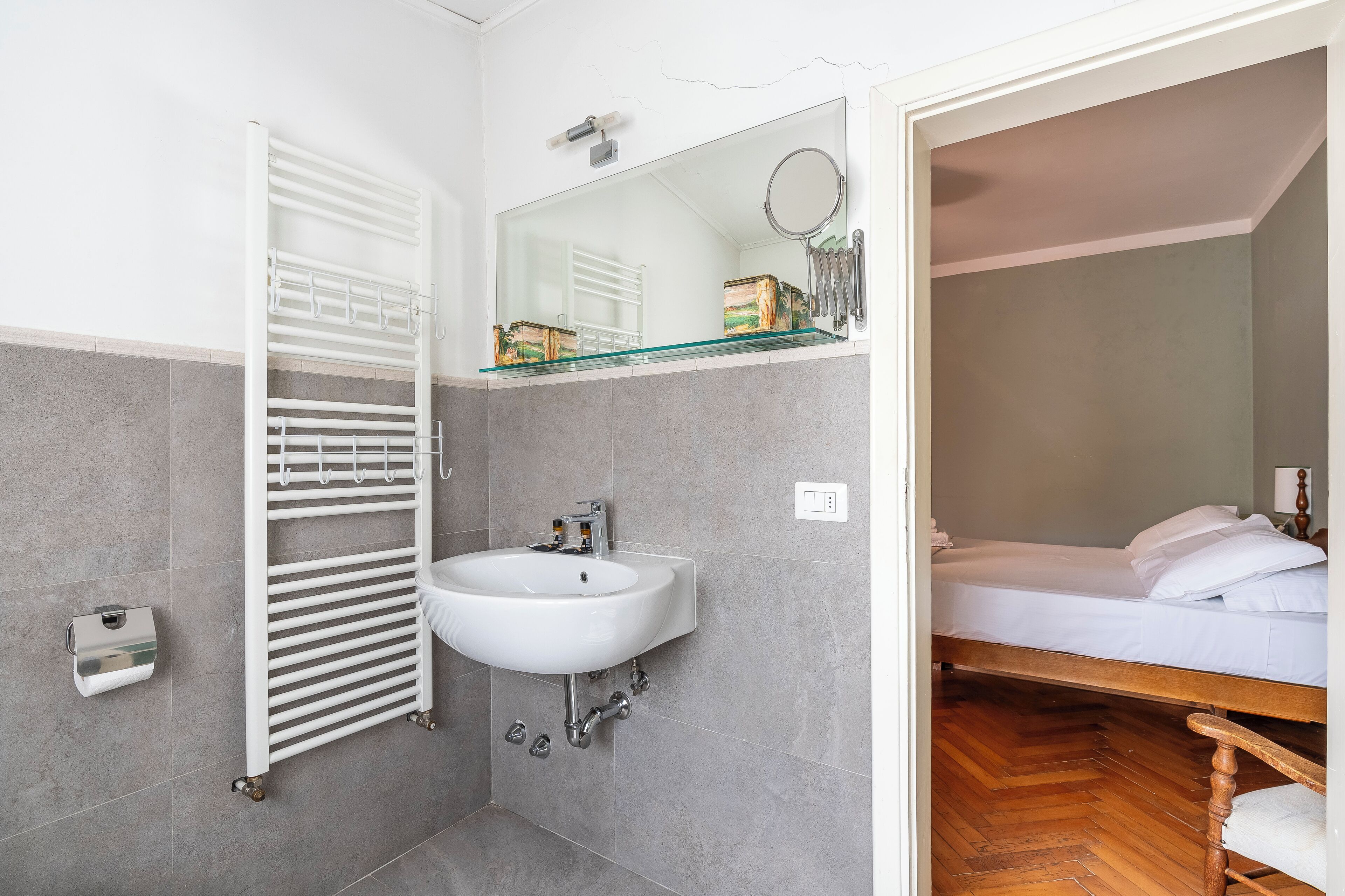 A bathroom with a white sink, a glass shelf, a towel rack, and a wooden bed in the adjacent room.