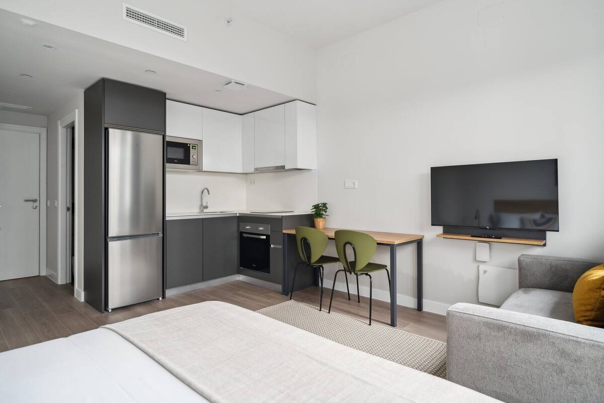 A modern kitchen with a microwave, sink, and stove, a dining area with chairs, a TV mounted on the wall, and a bed with a white comforter.
