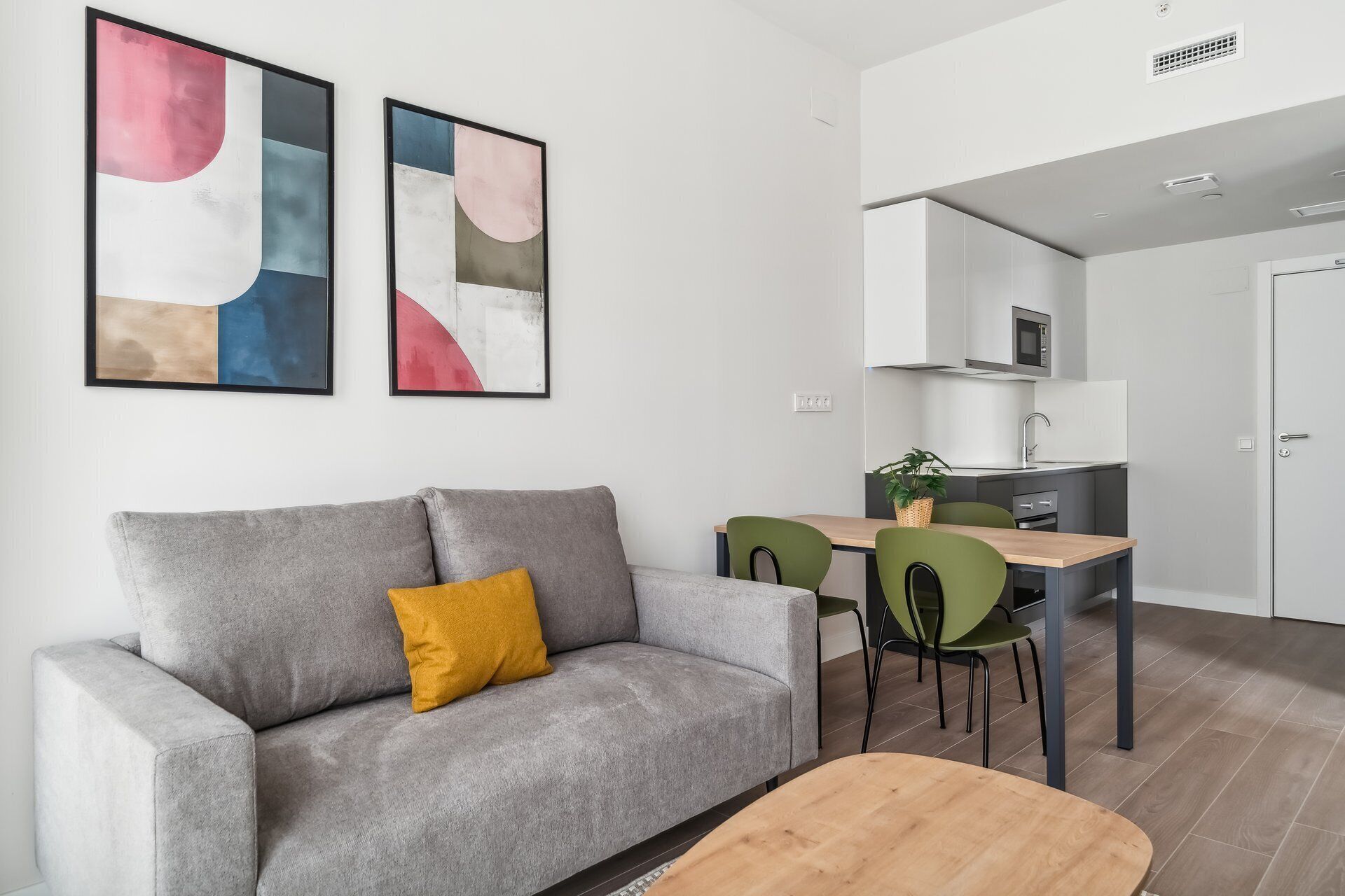 Un saló modern amb un sofà gris, una taula de centre de fusta i una zona de menjador amb cadires verdes. Hi ha quadres abstractes a la paret, un microones i un rentapeus a la cuina.