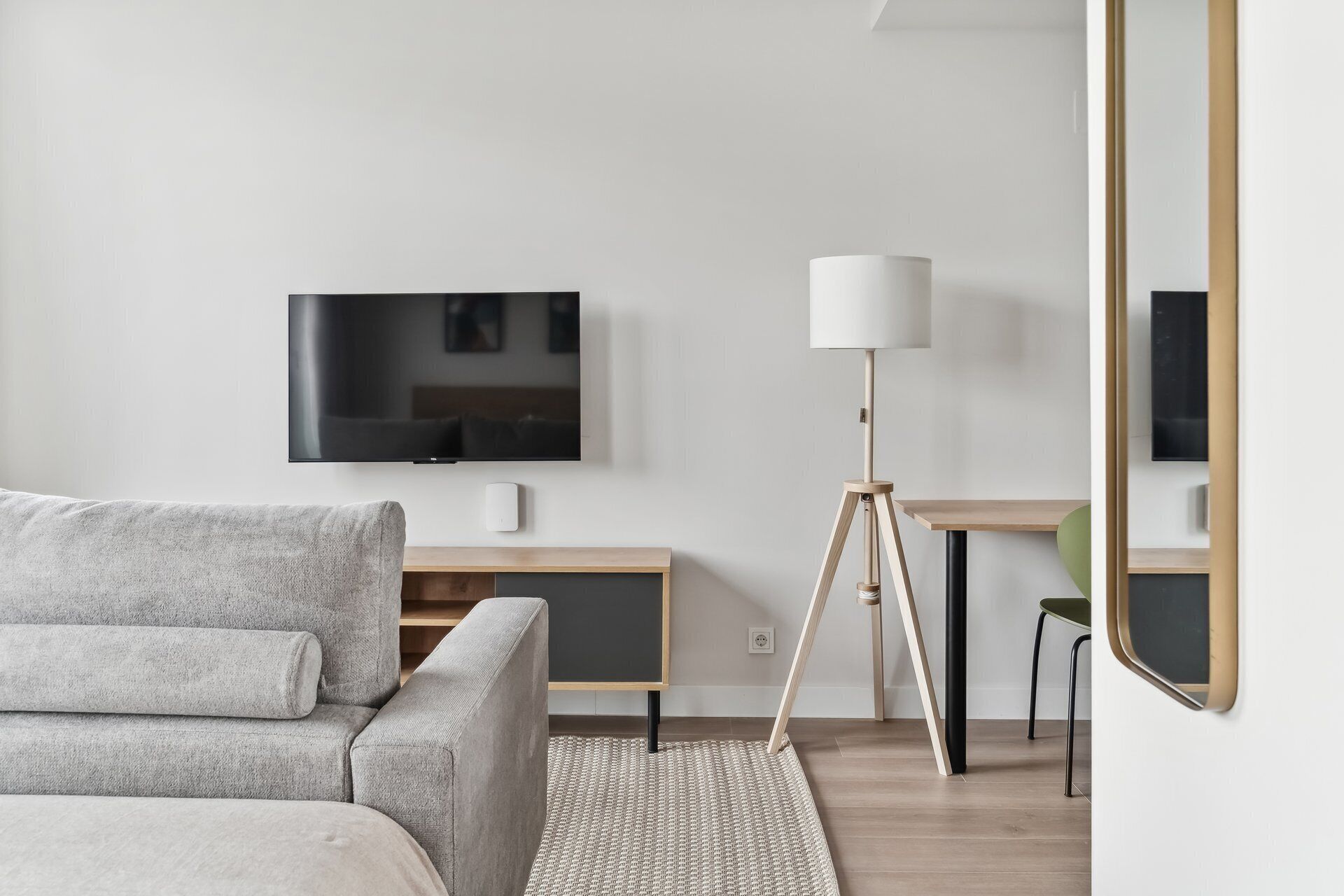 Una sala de estar moderna con un sofá gris, un televisor de pantalla plana montado en la pared, un escritorio de madera, una lámpara de pie y un espejo.
