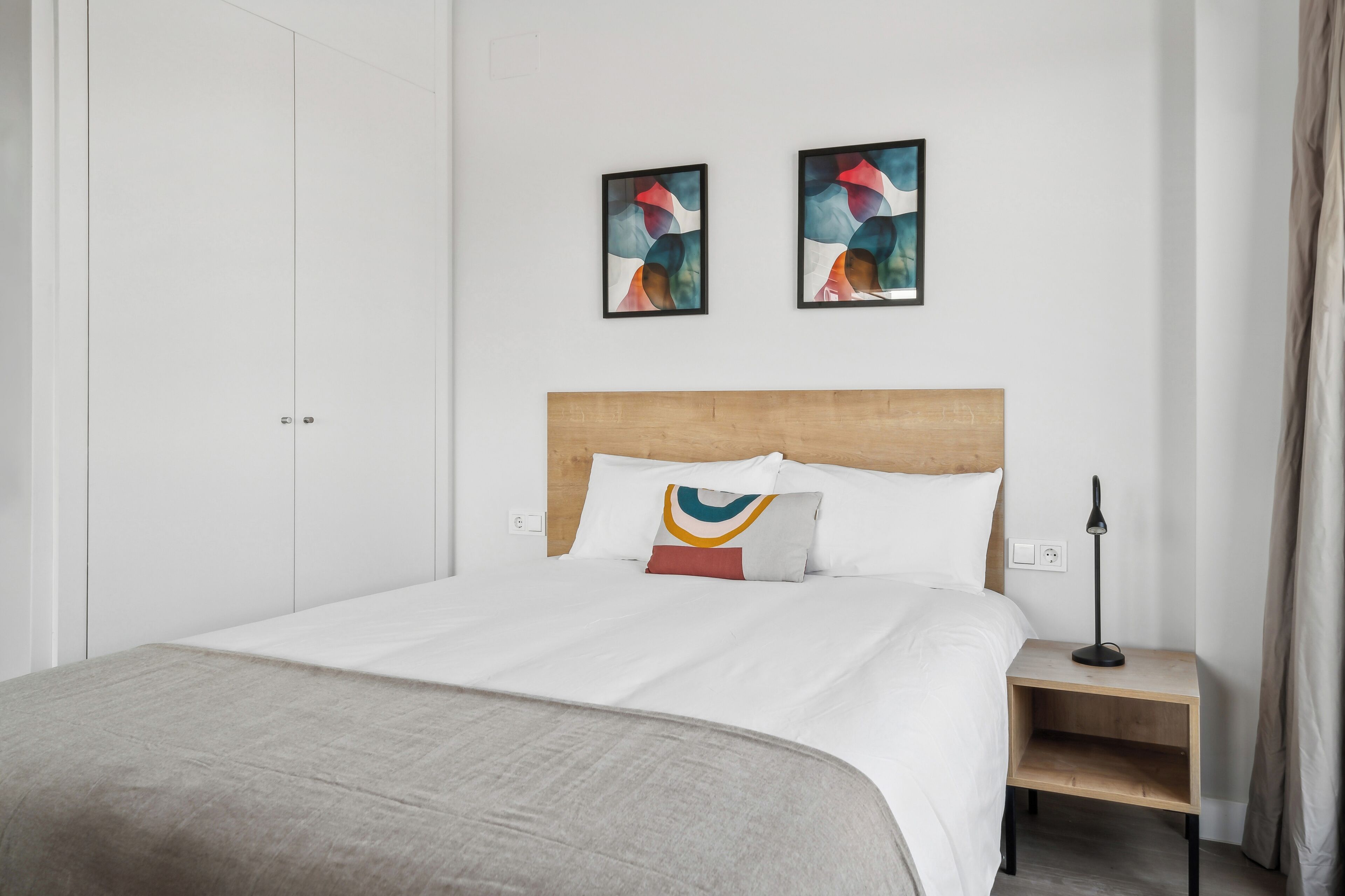 Quarto de hotel com cabeceira de madeira, cama com roupa de cama branca, duas pinturas abstratas na parede, mesa de cabeceira com luminária e um cobertor cinza.