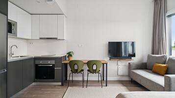 Una cocina moderna con horno empotrado, una zona de comedor con dos sillas, un televisor de pantalla plana y un sofá con un cojín mostaza.