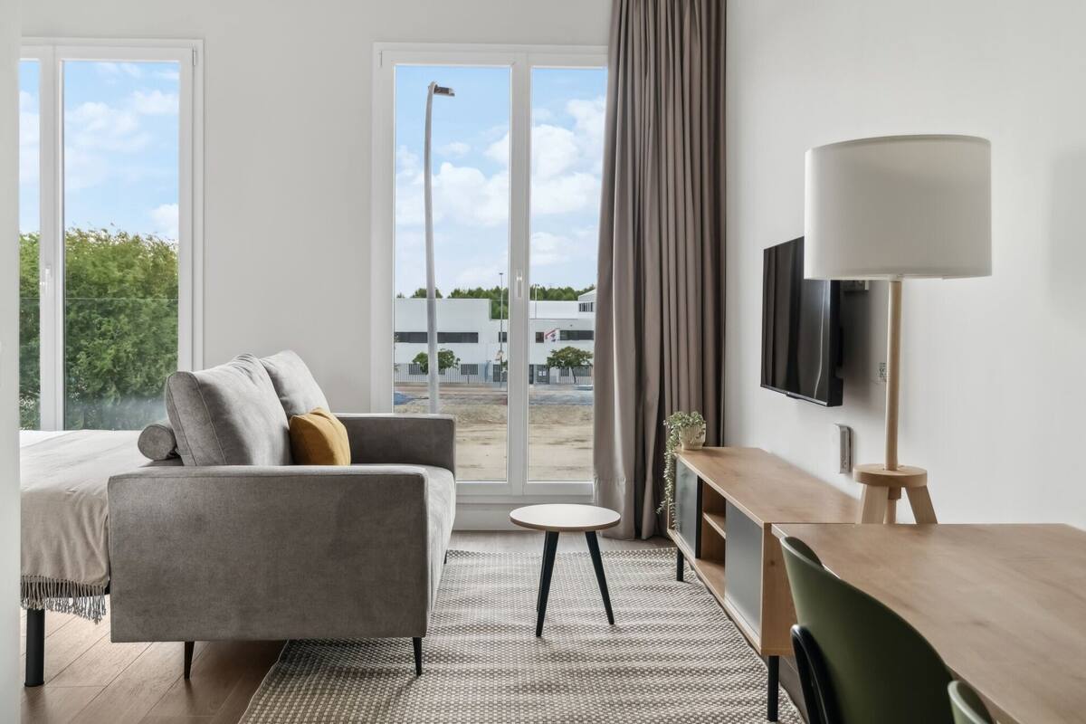 A modern hotel room with a sofa, a small round table, a TV mounted on the wall, and a large window with curtains.