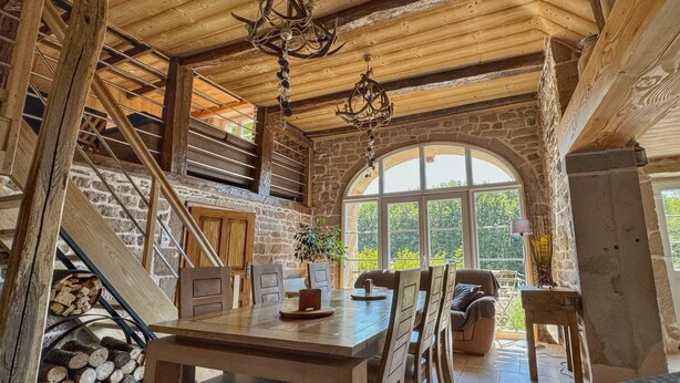 A rustic interior with a stone fireplace, wooden staircase, and a dining area with a view of greenery outside. - Maison Spacieuse , Pour Séjours au Calme sur le Plateau des Mille Étangs (La Lanterne-et-les-Armonts)