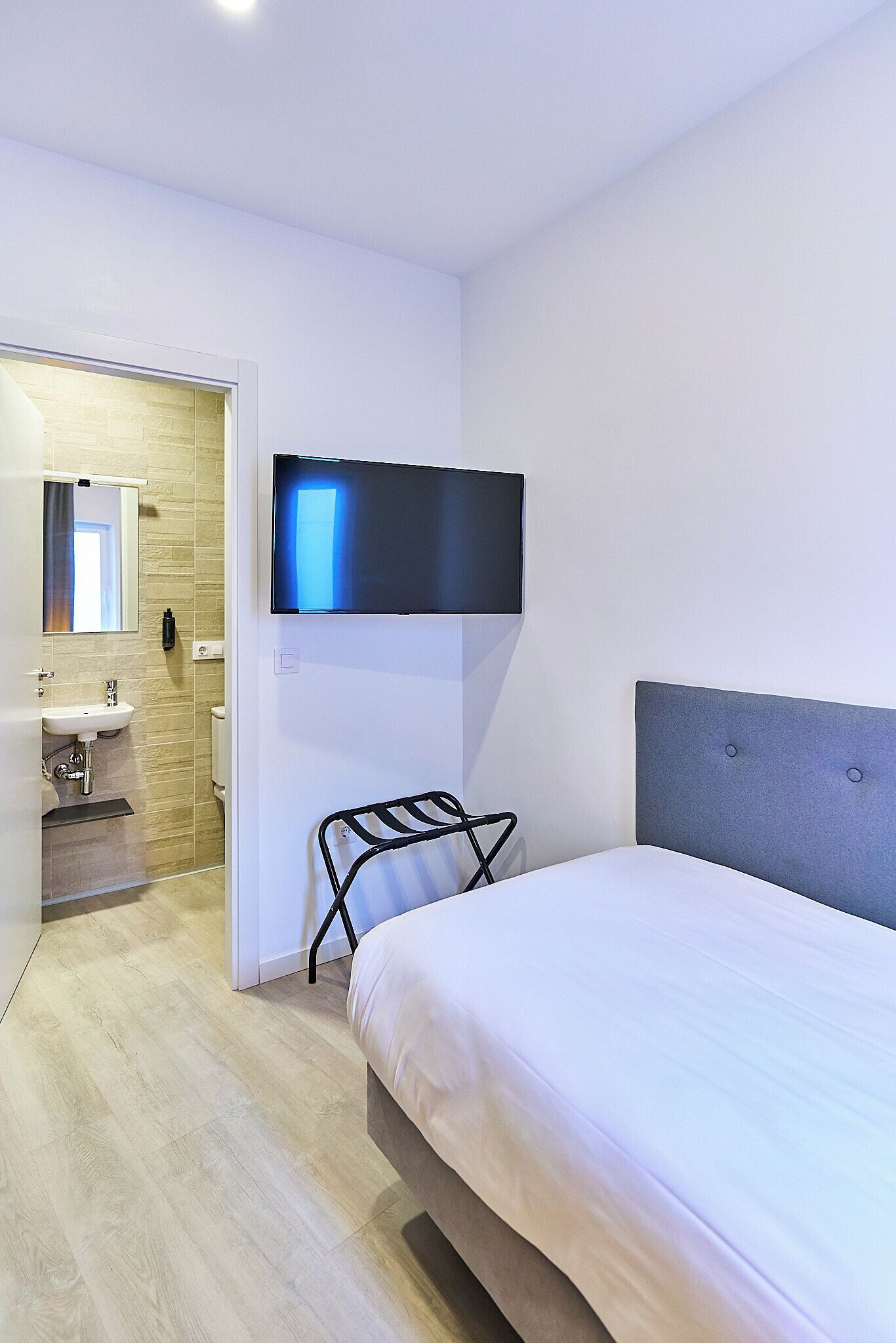 A modern hotel room with a bed, a mounted TV, a bathroom with a sink and toilet, and a wooden floor.