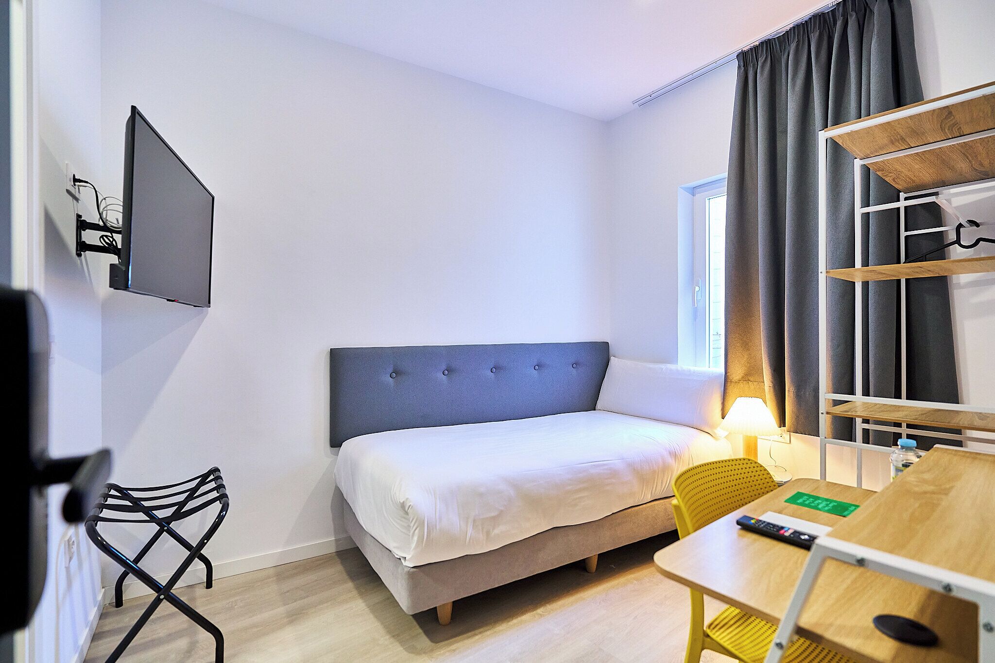 A hotel room with a bed, a desk, a chair, a TV mounted on the wall, and a window with curtains.