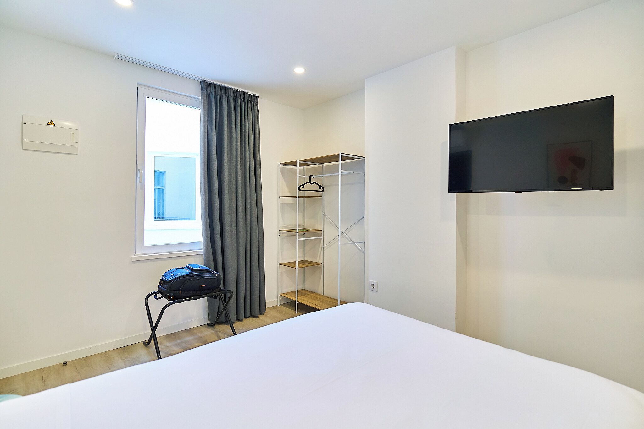 A bedroom with a bed, a TV mounted on the wall, a wardrobe, and a window with curtains.