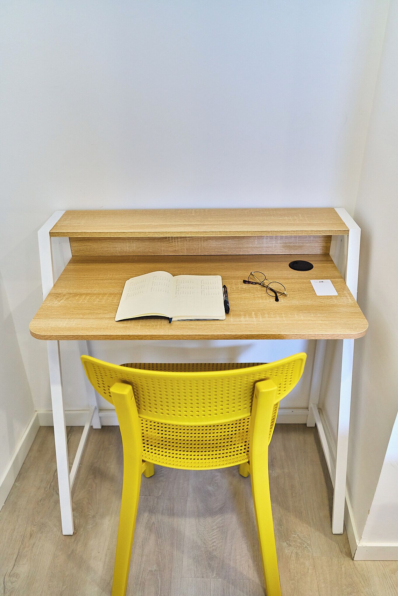 A wooden desk with a yellow chair, a notebook, glasses, and a pen.