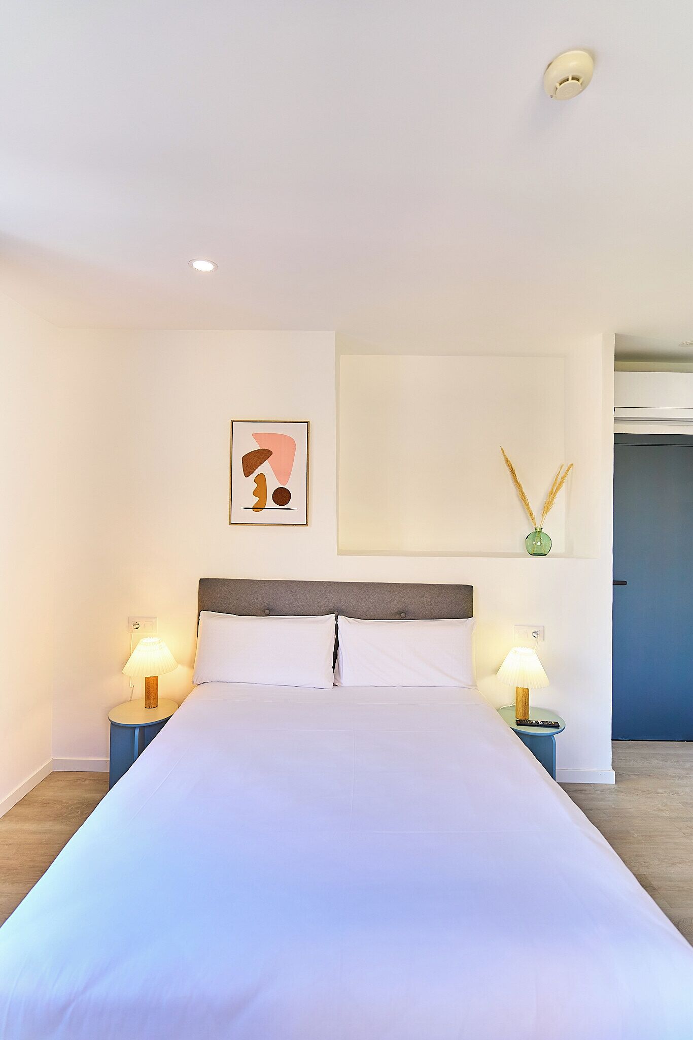 A bedroom with a large bed, two bedside tables with lamps, a wall-mounted artwork, and a small vase with decorative reeds.