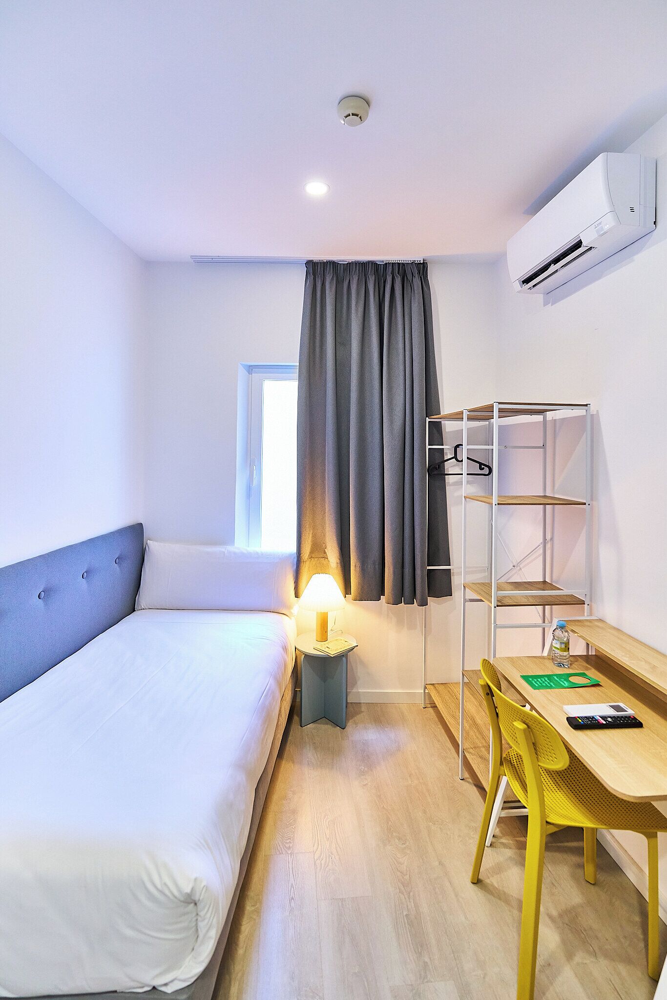 Habitació d'hotel amb un llit, una taula, una cadira, una làmpada, una estant i un aparell d'aire condicionat.