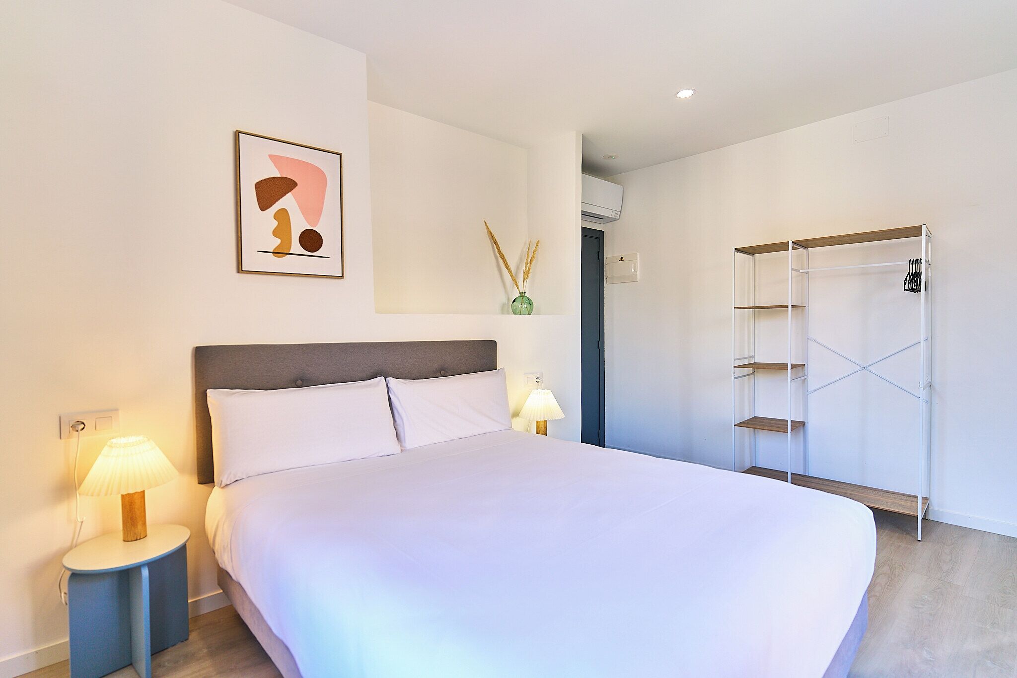 A neatly arranged hotel room with a large bed, bedside lamps, a wall-mounted shelf, and a wardrobe.