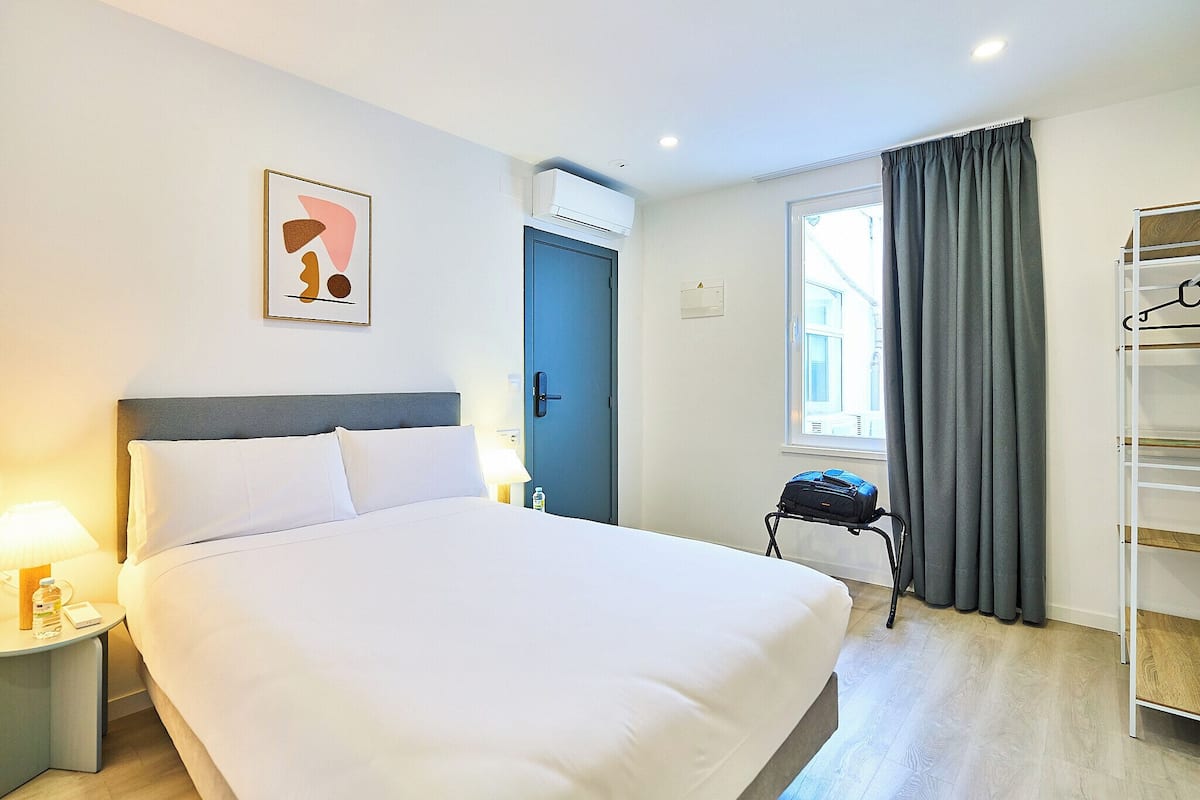 Habitación de hotel con cama, mesita de noche, lámpara, equipo de aire acondicionado, ventana con cortinas y perchero.