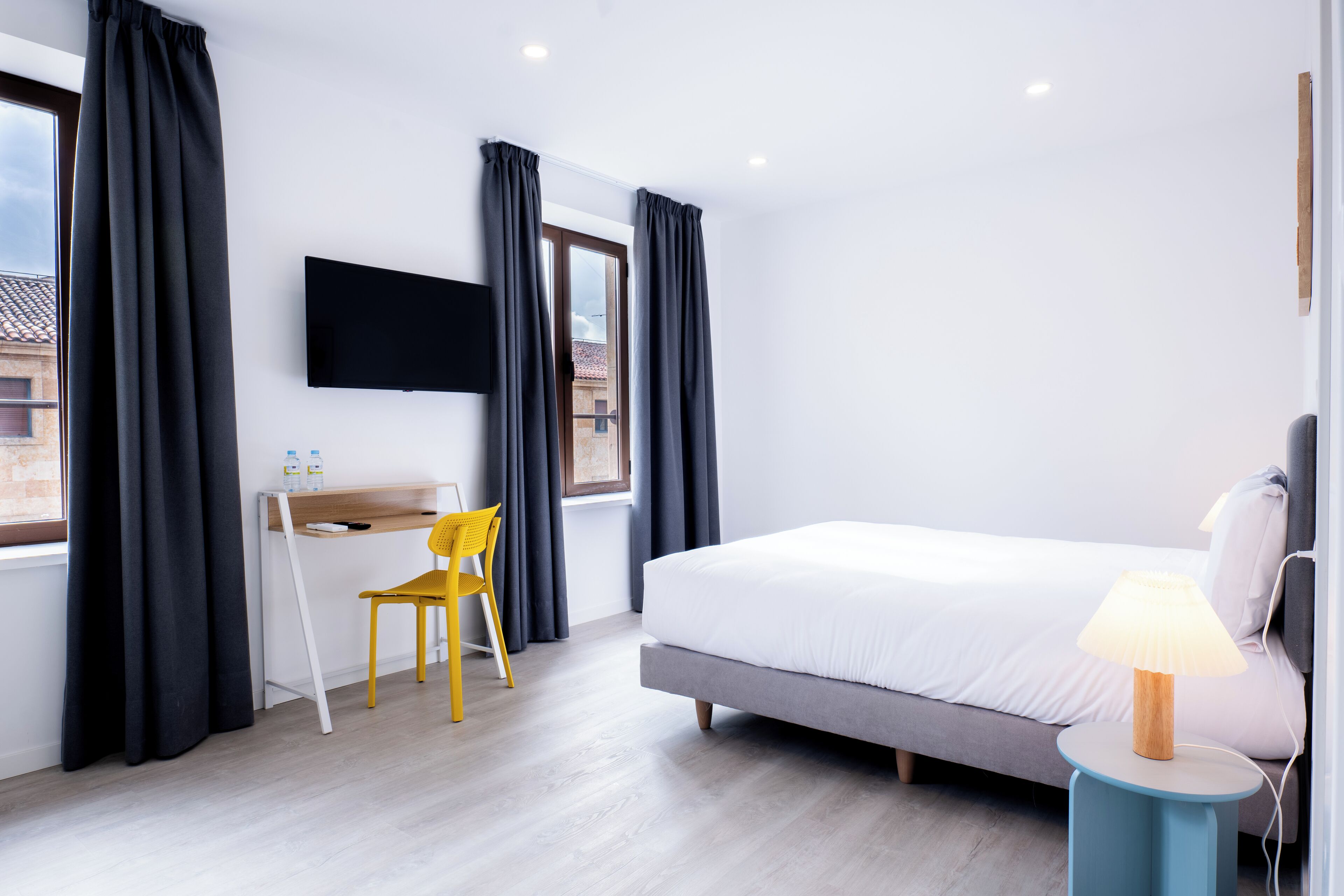 A modern hotel room with a bed, a desk, a chair, a TV, and a lamp.