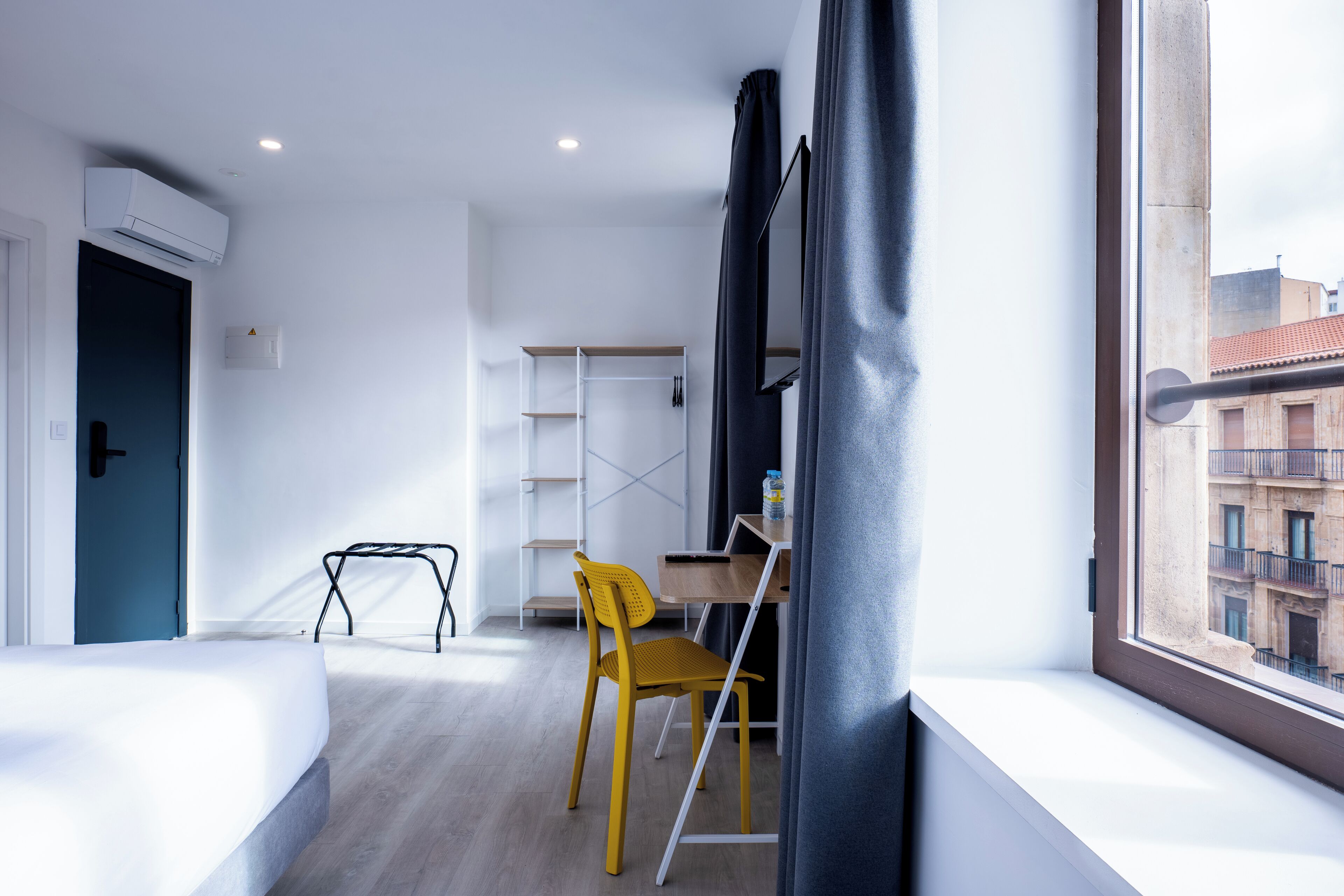 A modern hotel room with a bed, a desk, a chair, and a window with a view of buildings.