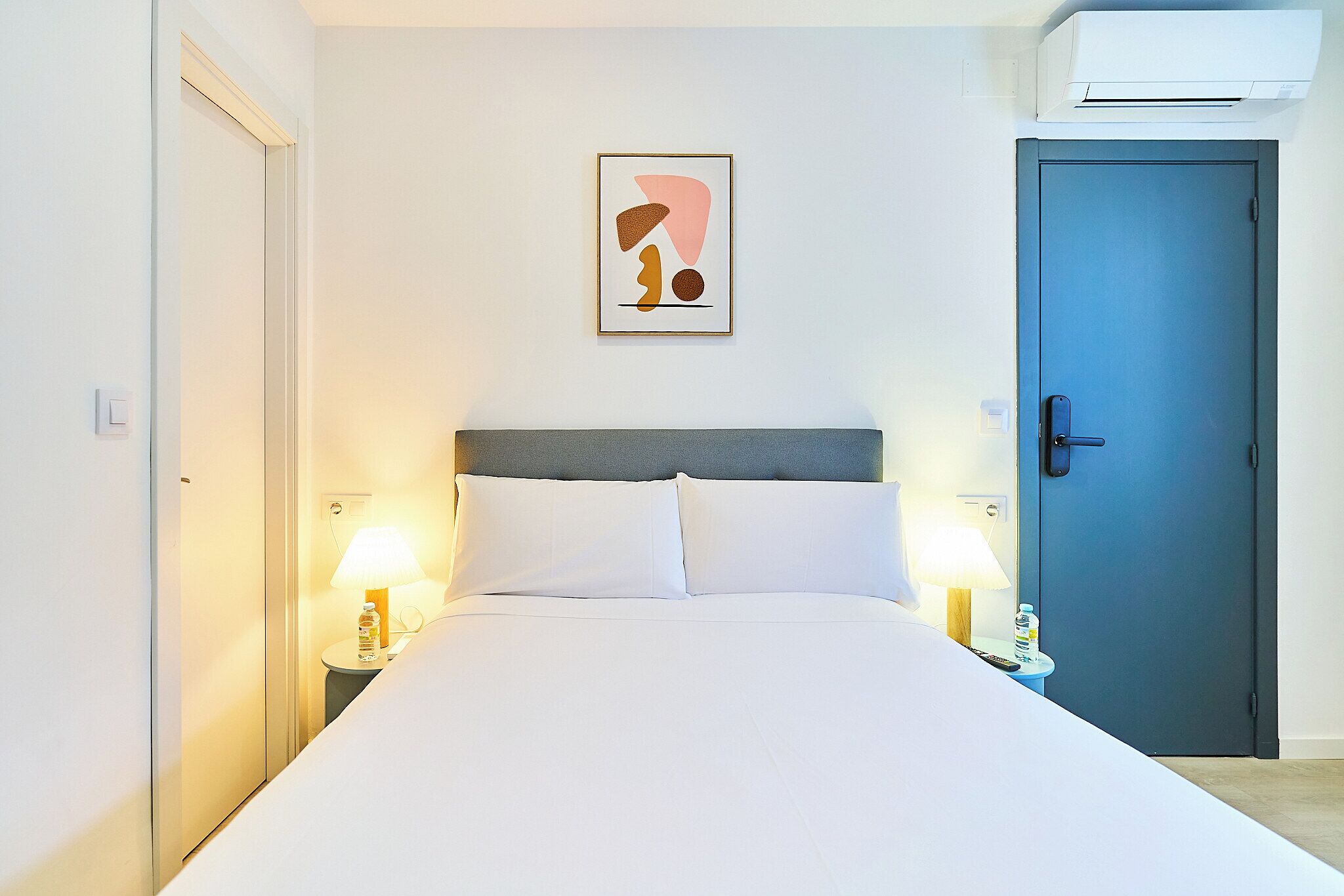 Una cama bien hecha con sábanas blancas, una obra de arte abstracta enmarcada encima, una mesita de noche con una lámpara y una puerta azul.