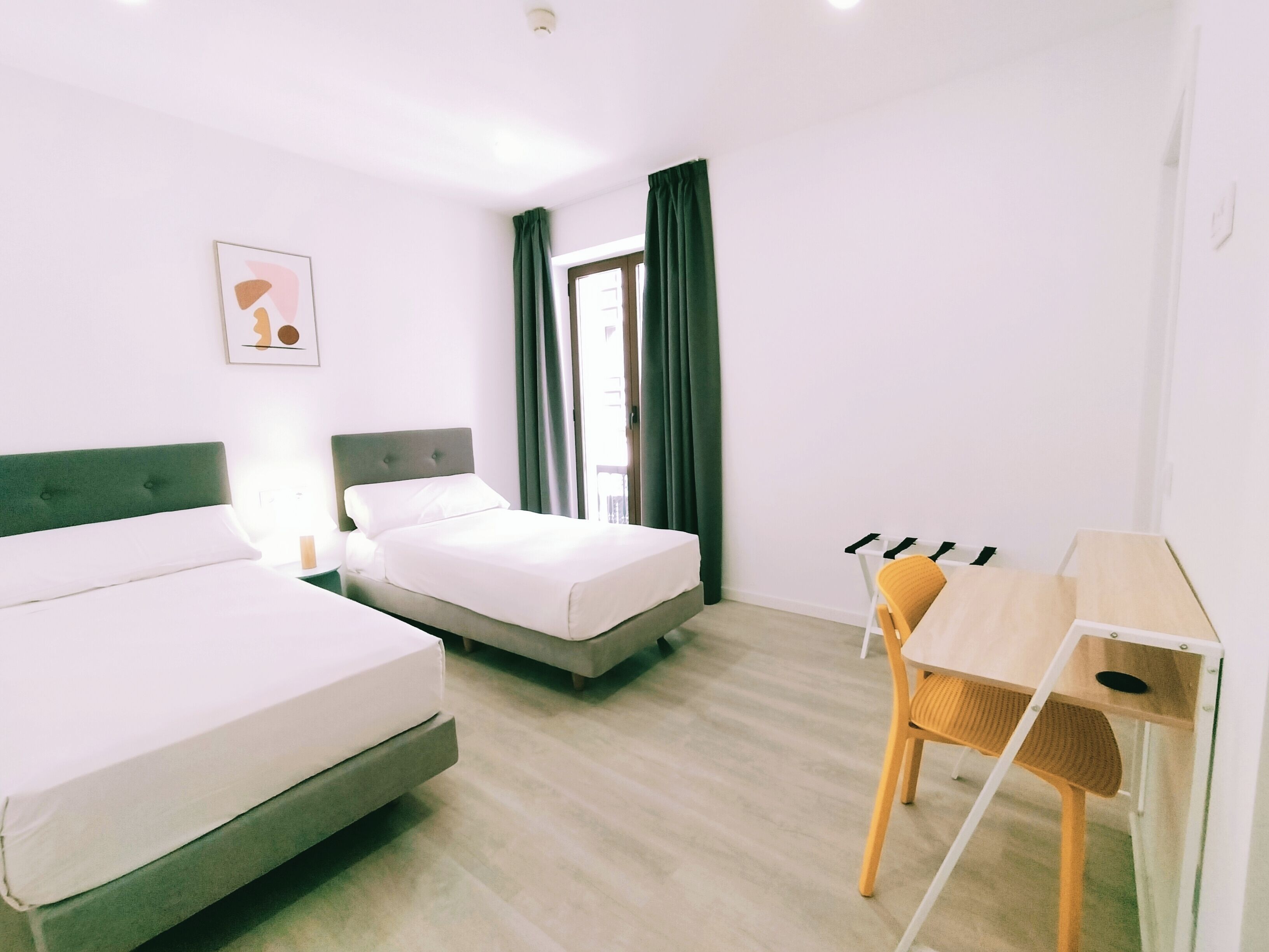 Habitación de hotel con dos camas individuales, un escritorio de madera, una silla y una ventana con cortinas.