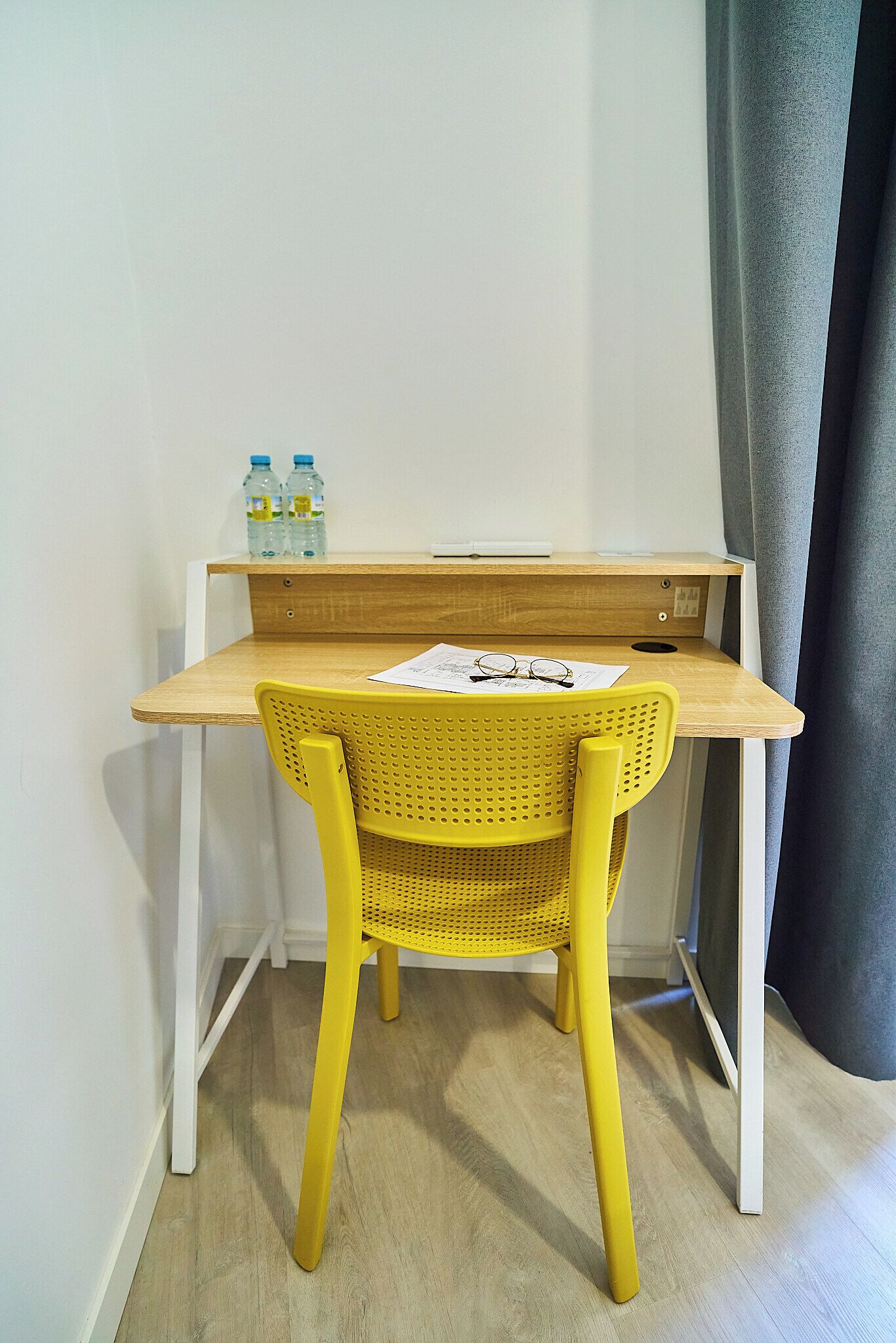 A small wooden desk with a yellow chair, a pair of glasses, and two water bottles on top.