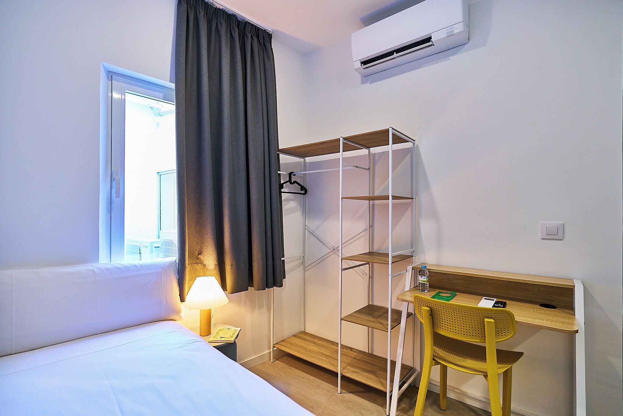 A hotel room with a bed, a desk with a chair, a shelf unit, a lamp, and a window with curtains.