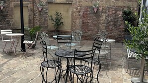 An outdoor patio with stone flooring, metal tables and chairs, potted plants, and a stone wall with windows.