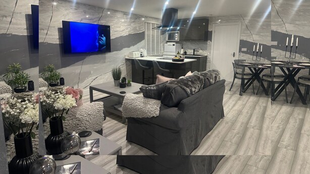 A modern living room with a grey sofa, a coffee table, and a TV. There's a kitchen area with a dining table and chairs.