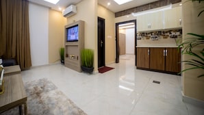 A modern living room with a flat-screen TV, a sofa, and a coffee table. There is a kitchen area with wooden cabinets and a countertop.