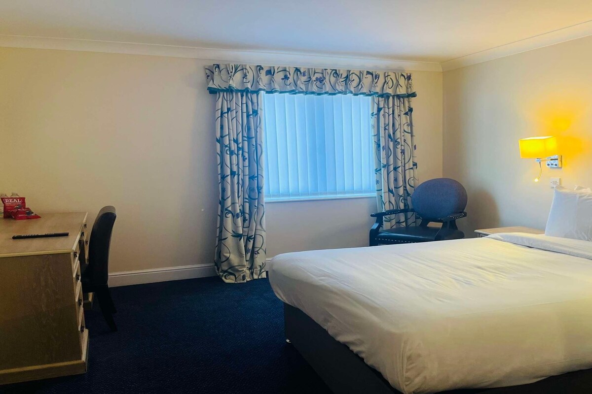 Une chambre d’hôtel avec un lit, un bureau, une chaise, une lampe et une fenêtre avec des rideaux.