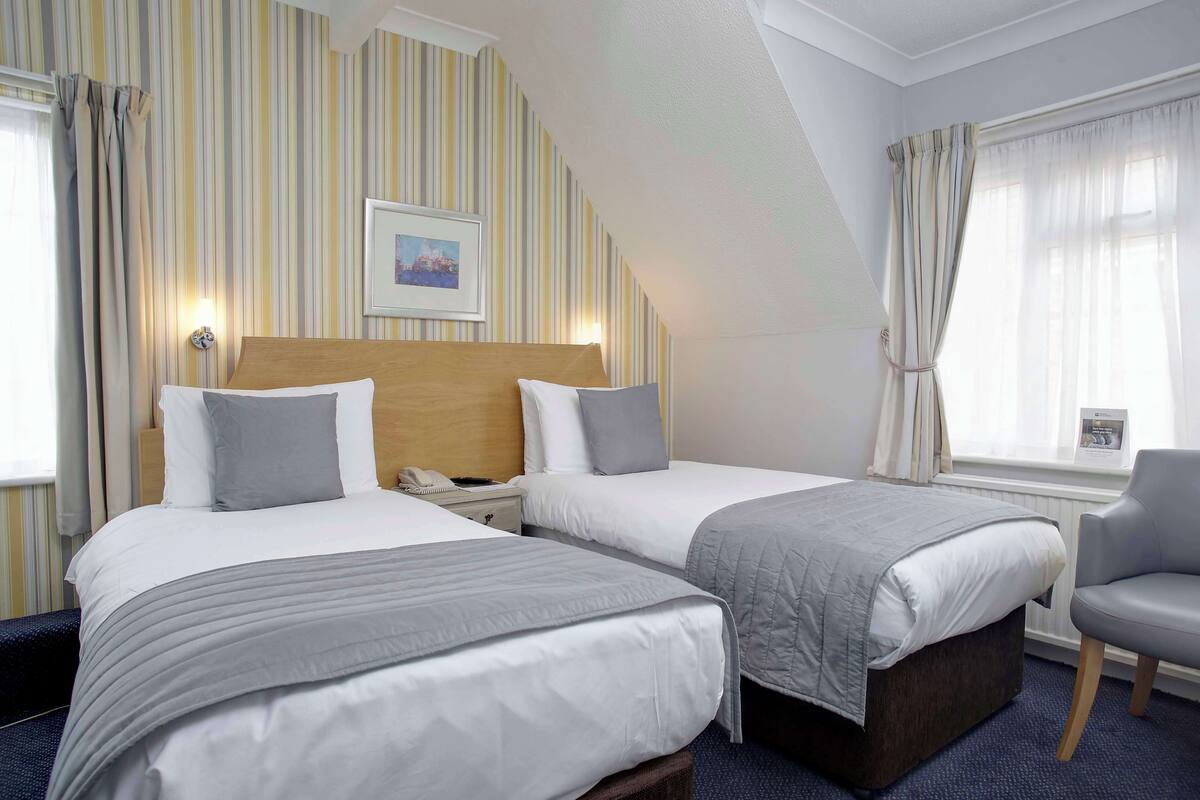 Une chambre d’hôtel avec deux lits, une tête de lit en bois, une table de chevet, une chaise et une fenêtre avec des rideaux.