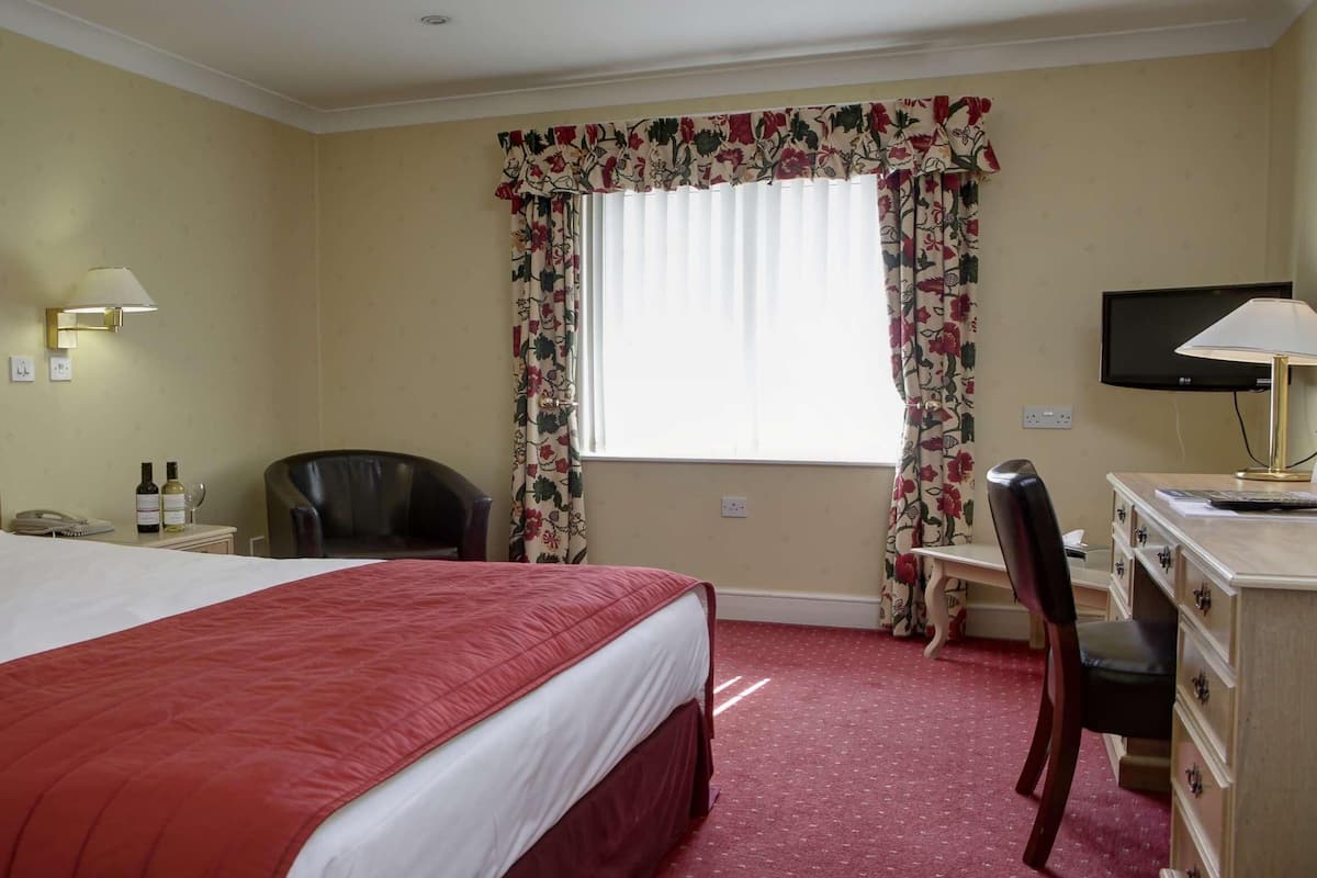 Une chambre d’hôtel avec un grand lit, un bureau avec une chaise, une télévision et une fenêtre agrémentée de rideaux à motifs floraux.
