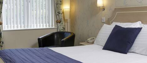 A hotel room with a bed, a chair, a bedside table with a lamp, and a window with blinds.