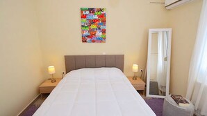 Ein ordentlich bezogenes Bett mit weißer Daunendecke, zwei Nachttische mit Lampen, ein farbenfrohes abstraktes Kunstwerk an der Wand und ein Fenster mit Vorhängen.