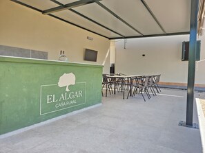 A green wall with 'EL ALGAR CASA RURAL' signage, a covered outdoor seating area with tables and chairs, and a concrete floor.