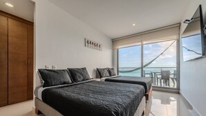 A modern bedroom with two beds, a large wardrobe, a flat-screen TV, and a balcony with a view of the sea.