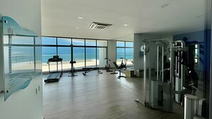 A gym with exercise equipment and large windows overlooking the beach.