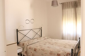 Een slaapkamer met een bed, nachtkastje, lamp en raam met gordijnen.
